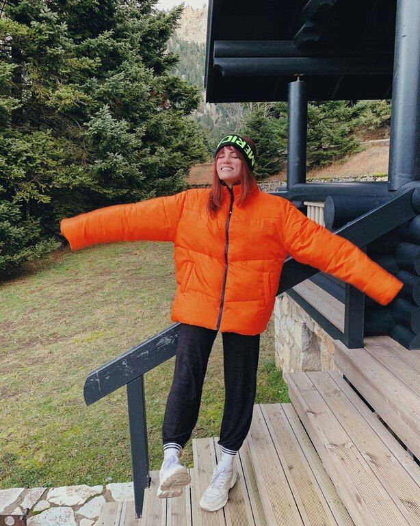 Η Μαίρη Συνατσάκη με πορτοκαλί φλούο puffer jacket, μαύρο παντελόνι και sneakers.