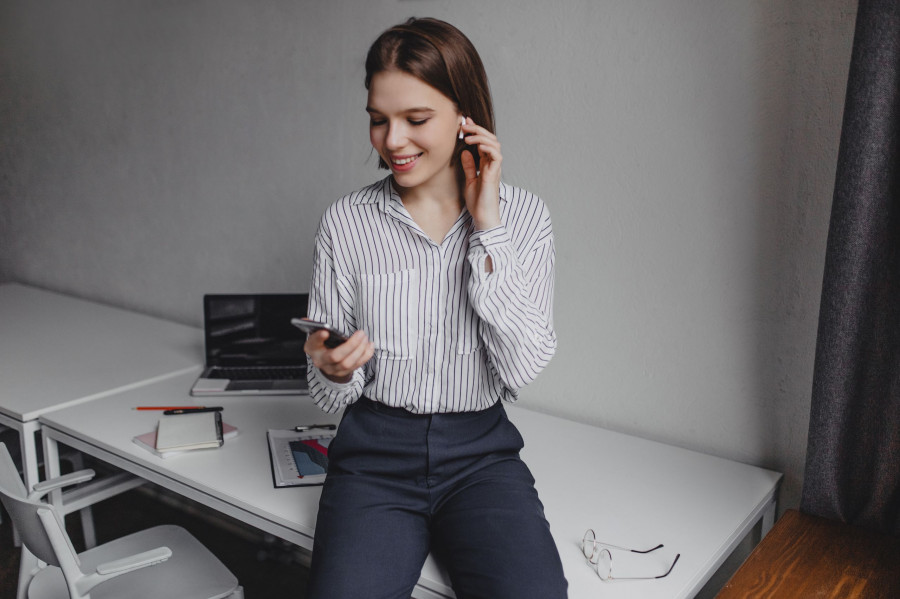 Charming woman in black pants and light blouse is sitting on table. Office worker listens to music